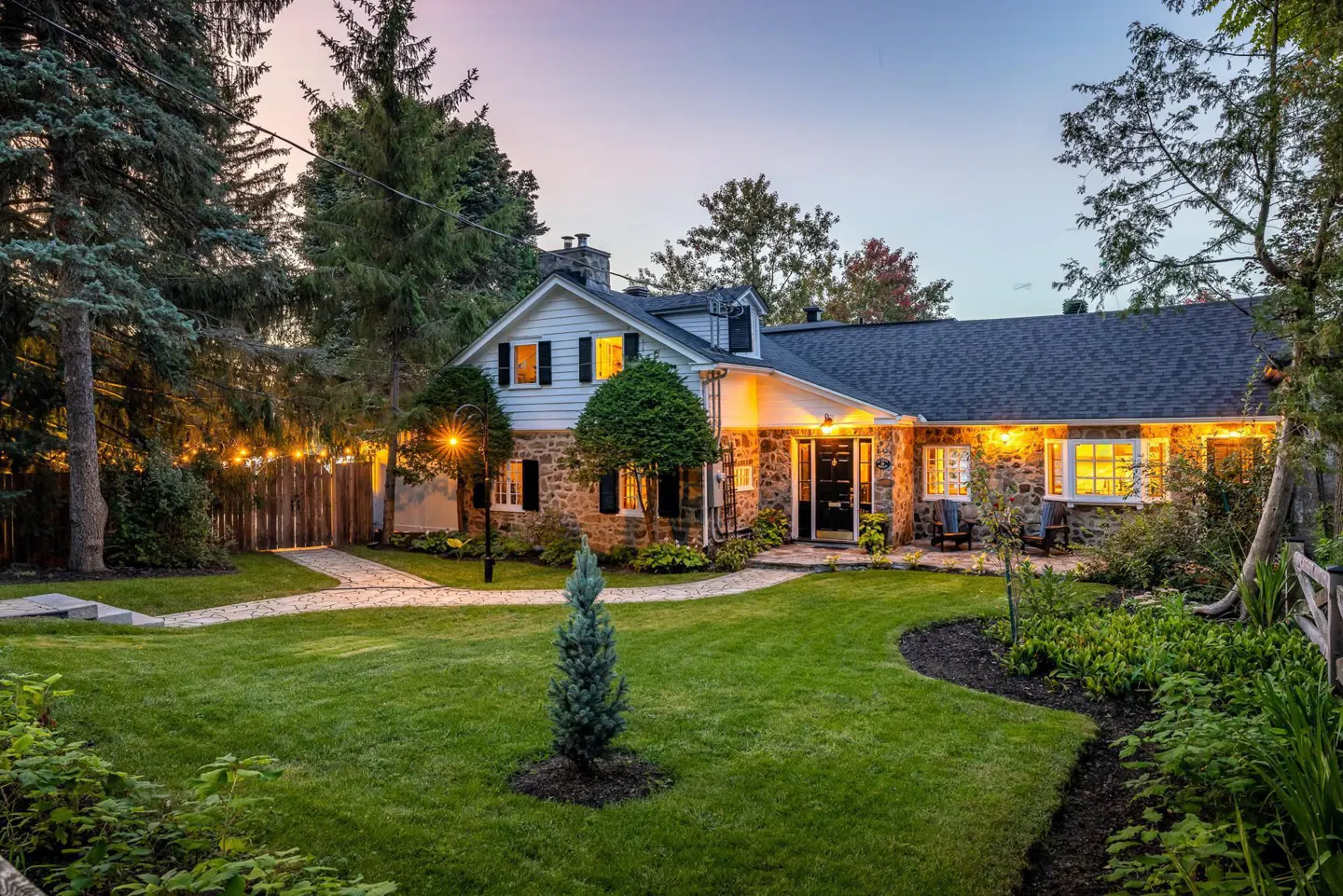 A charming stone house with a manicured lawn at dusk. Lights illuminate the windows and a stone path leading to the front door.