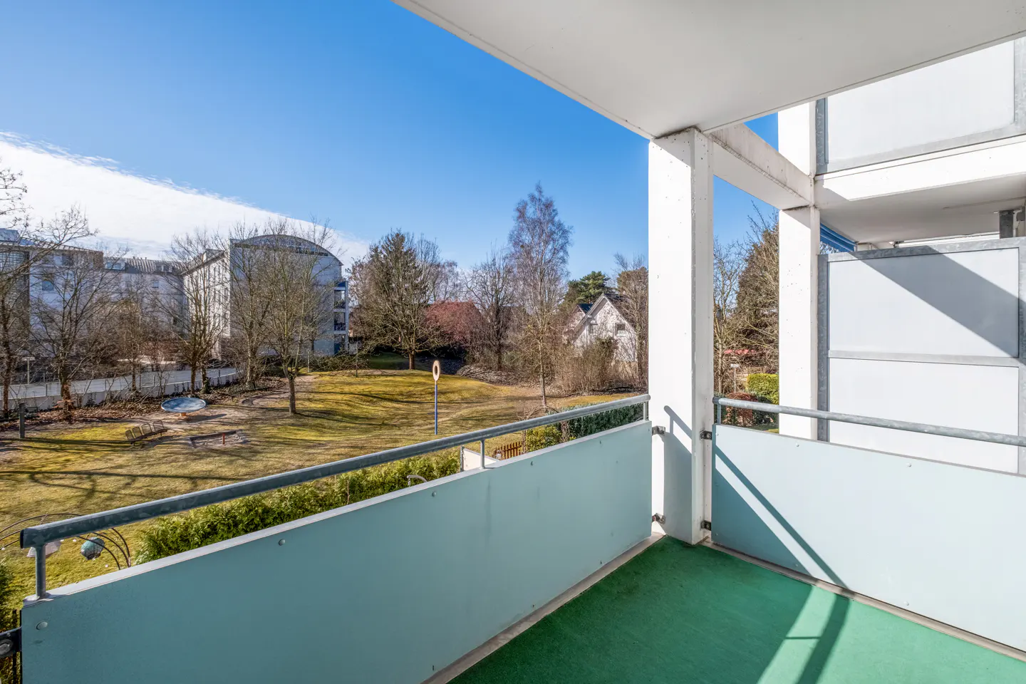 View from a balcony with green flooring, a light blue railing, and white support beams, overlooking a grassy yard with trees and buildings.