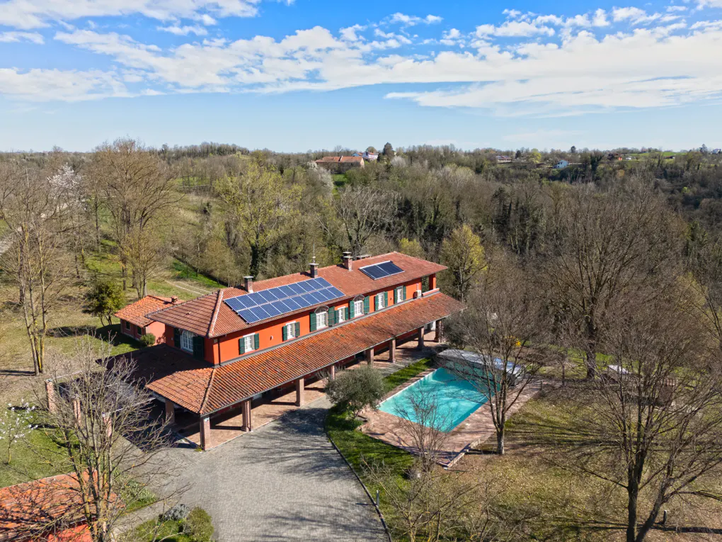 Aerial view of a red house with solar panels, a pool, and a tile roof surrounded by trees under a blue sky.