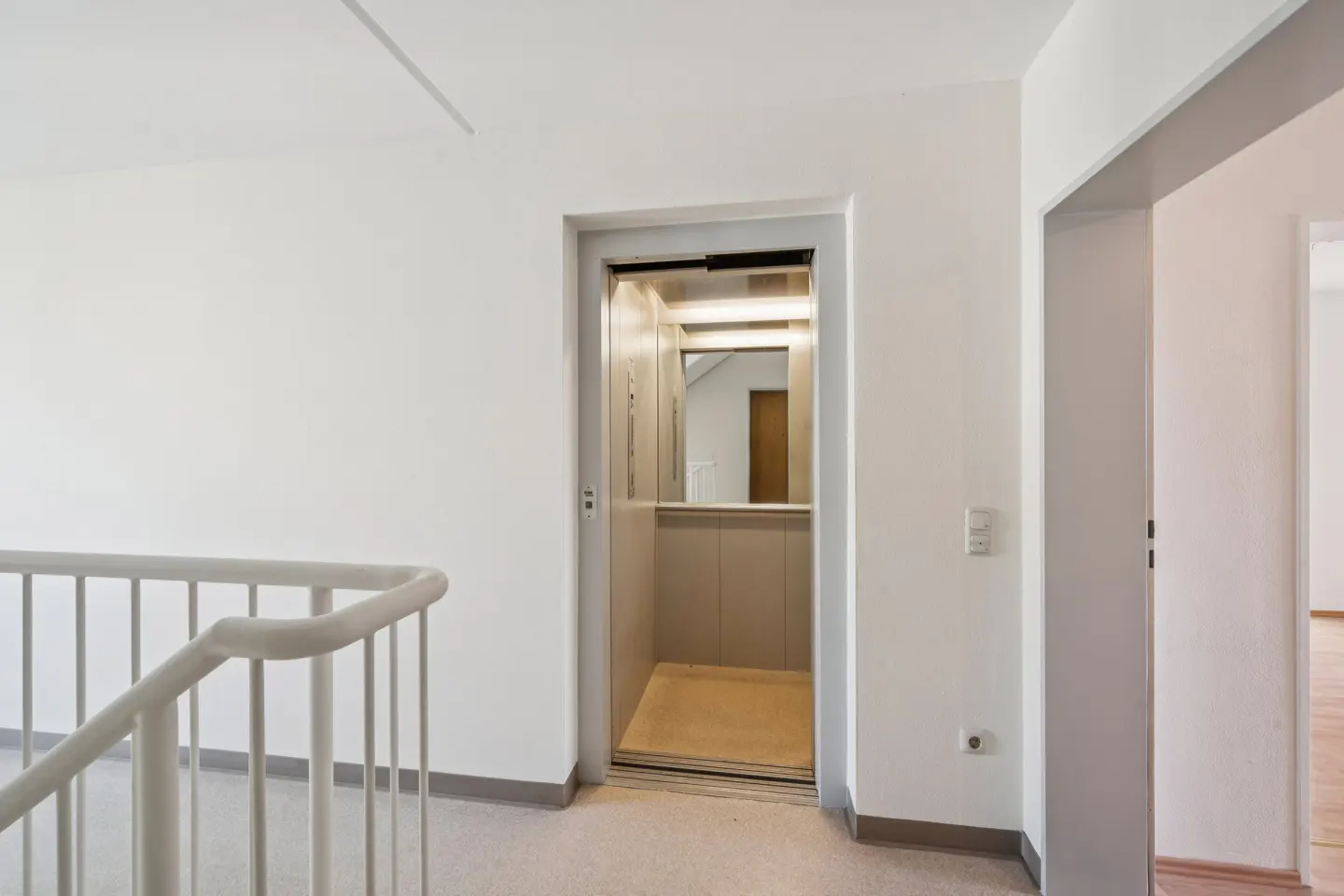 Interior view of a building with an open elevator, white walls, and a white staircase railing.