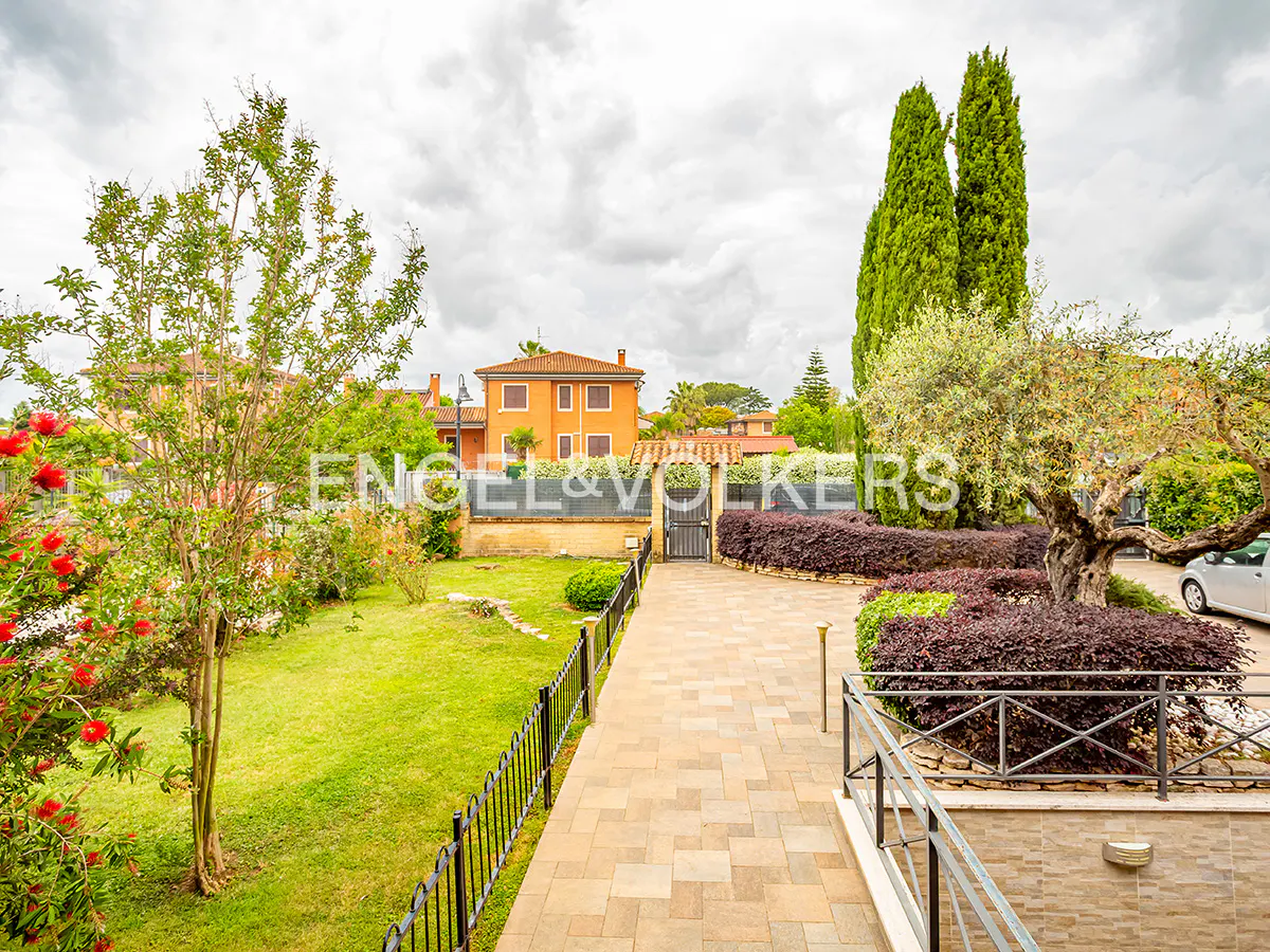 Exterior view of a property with a brick walkway, green lawn, trees, and an orange house in the background under a cloudy sky.