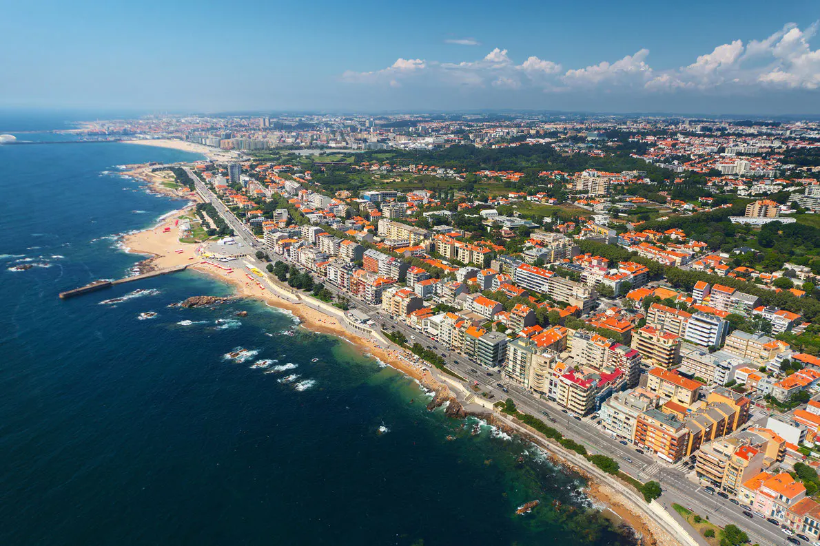 Aerial view of a coastal city with orange-roofed buildings, sandy beaches, and blue ocean under a partly cloudy sky.
