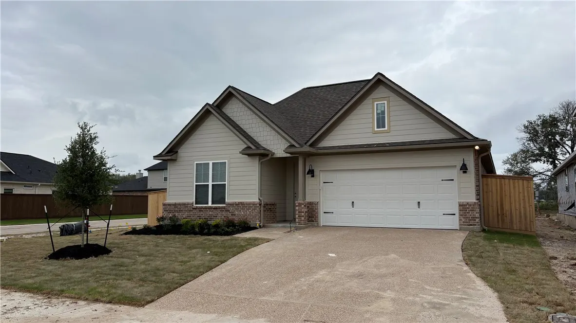 Beige single-story house with a brown roof, white garage door, and a small tree in the front yard.