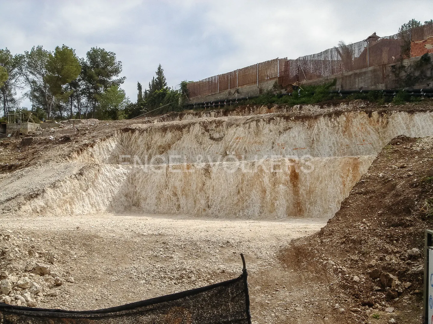 Construction site with a large excavated area, beige soil, trees, and a fence in the background.