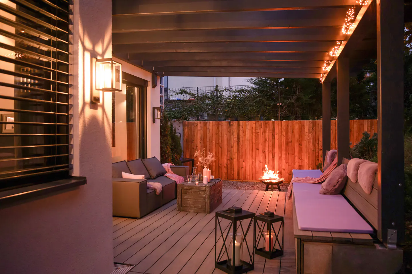 Cozy outdoor patio at dusk with a fire pit, modern lounge seating, string lights, and lanterns on a wooden deck.