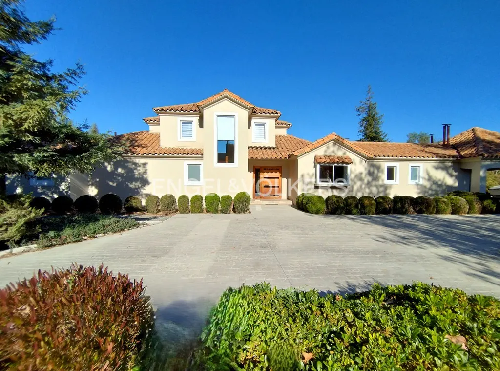 Beige two-story house with a red tile roof, a wooden front door, and a concrete driveway on a sunny day.