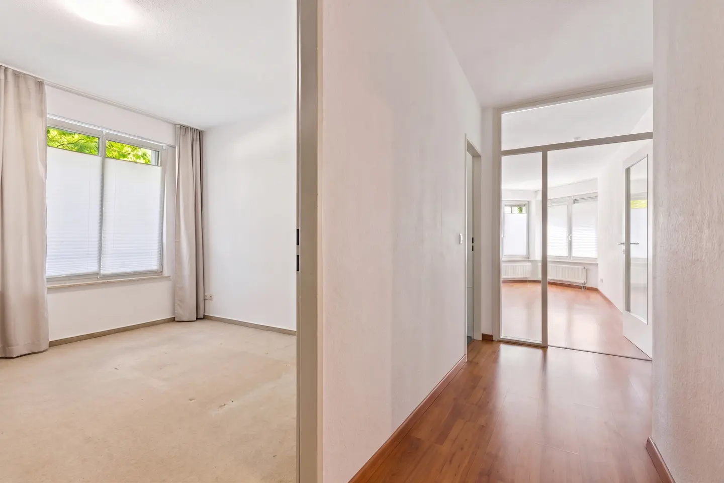 Hallway view into rooms. One room has beige carpet, white walls, window with blinds and curtains. The other has wood floors and glass doors.
