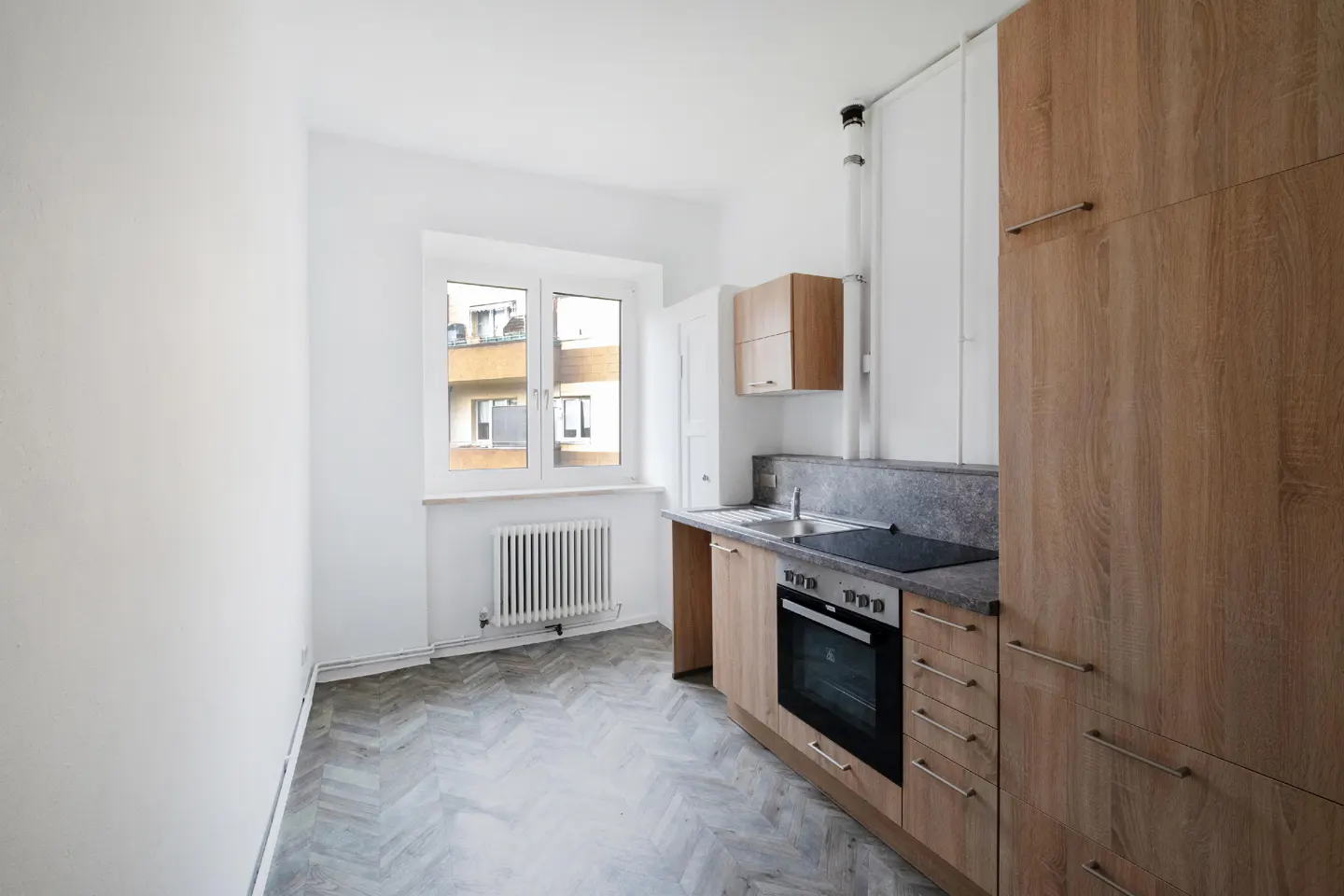 Bright kitchen with wood cabinets, gray countertops, and herringbone floors. A window overlooks a building.