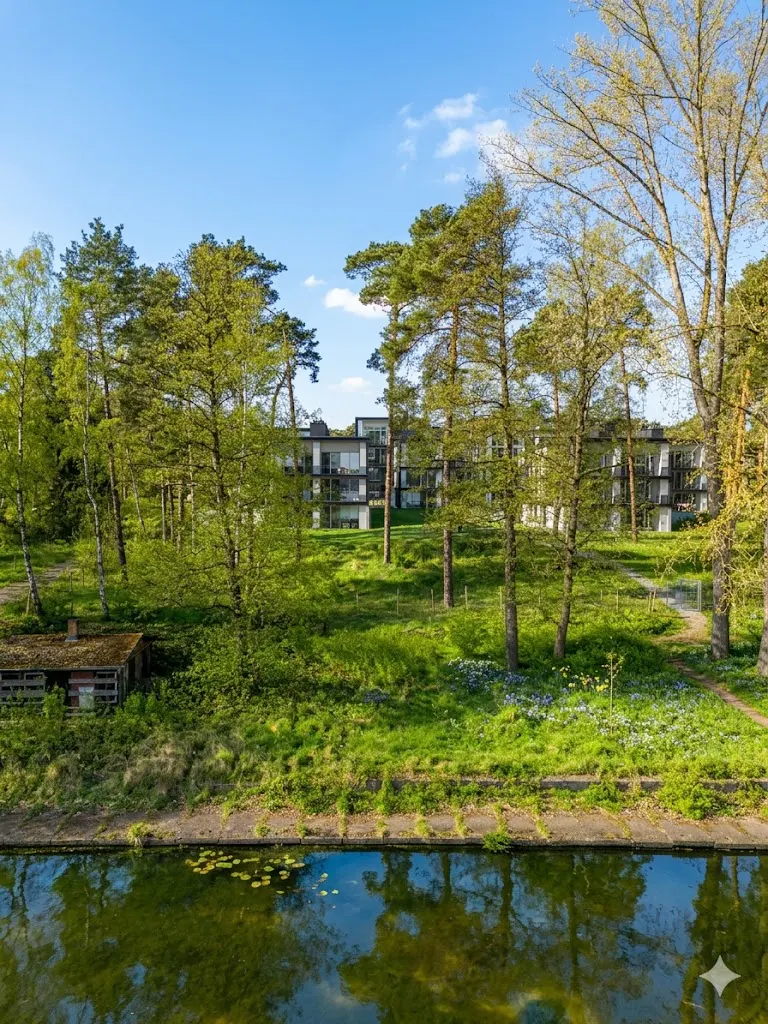 Apartment building seen through trees, reflected in a canal. Green grass and blue sky.