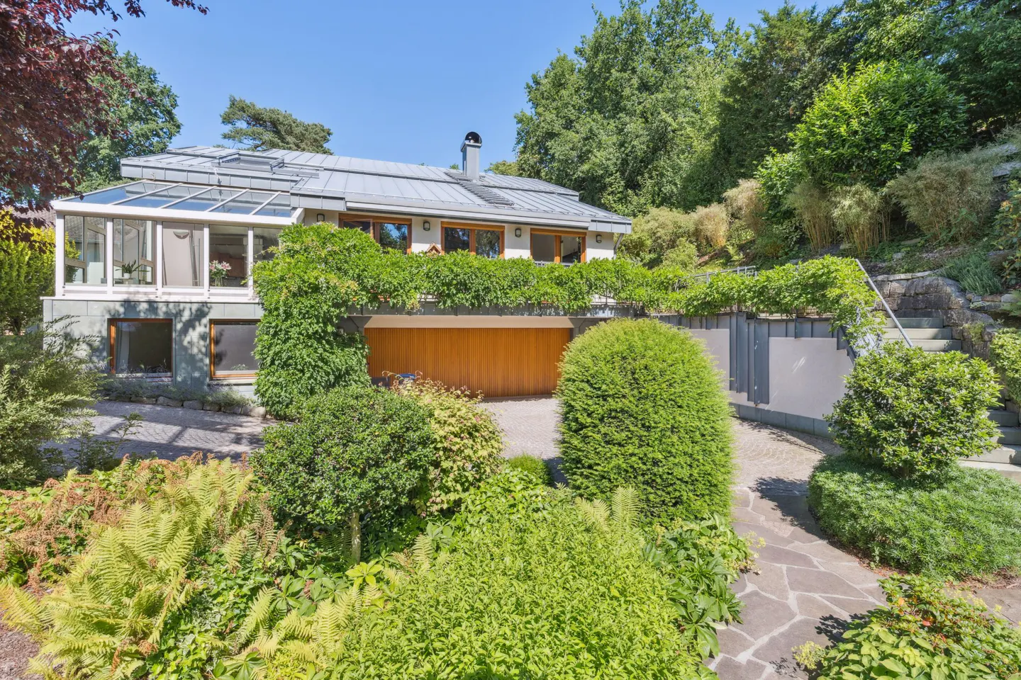 Exterior view of a modern home with a gray roof, wooden garage door, and lush green landscaping. Stone pathway leads to the entrance.