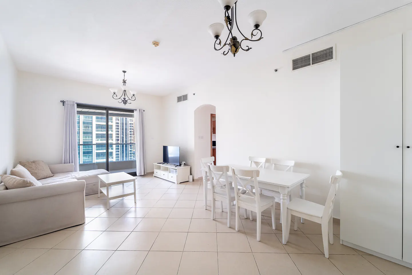 Bright, open-plan apartment with white walls, tile floors, and modern furnishings. A beige sofa faces a balcony with city views. A white dining set sits nearby.