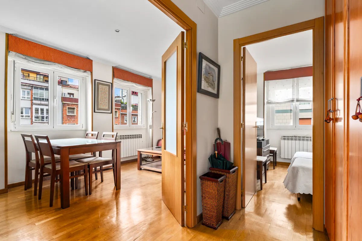 Bright apartment interior with wood floors, dining table, and open doorway to a bedroom. Windows overlook city buildings.