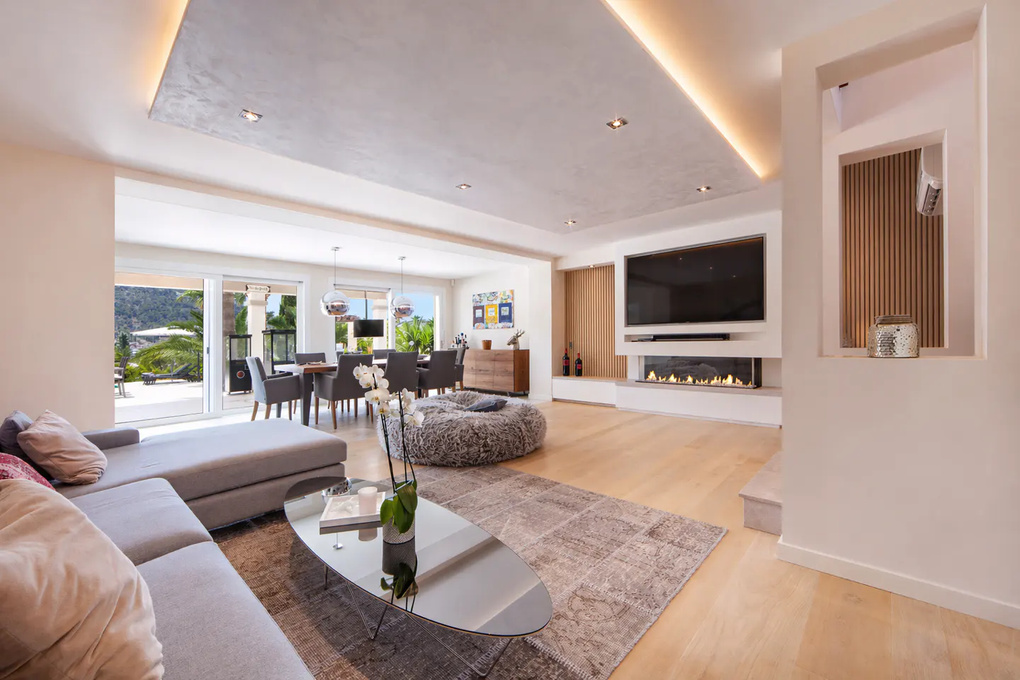 Bright, open living room with a gray sectional, glass table, and fireplace with a TV above it. Dining area visible through sliding glass doors.
