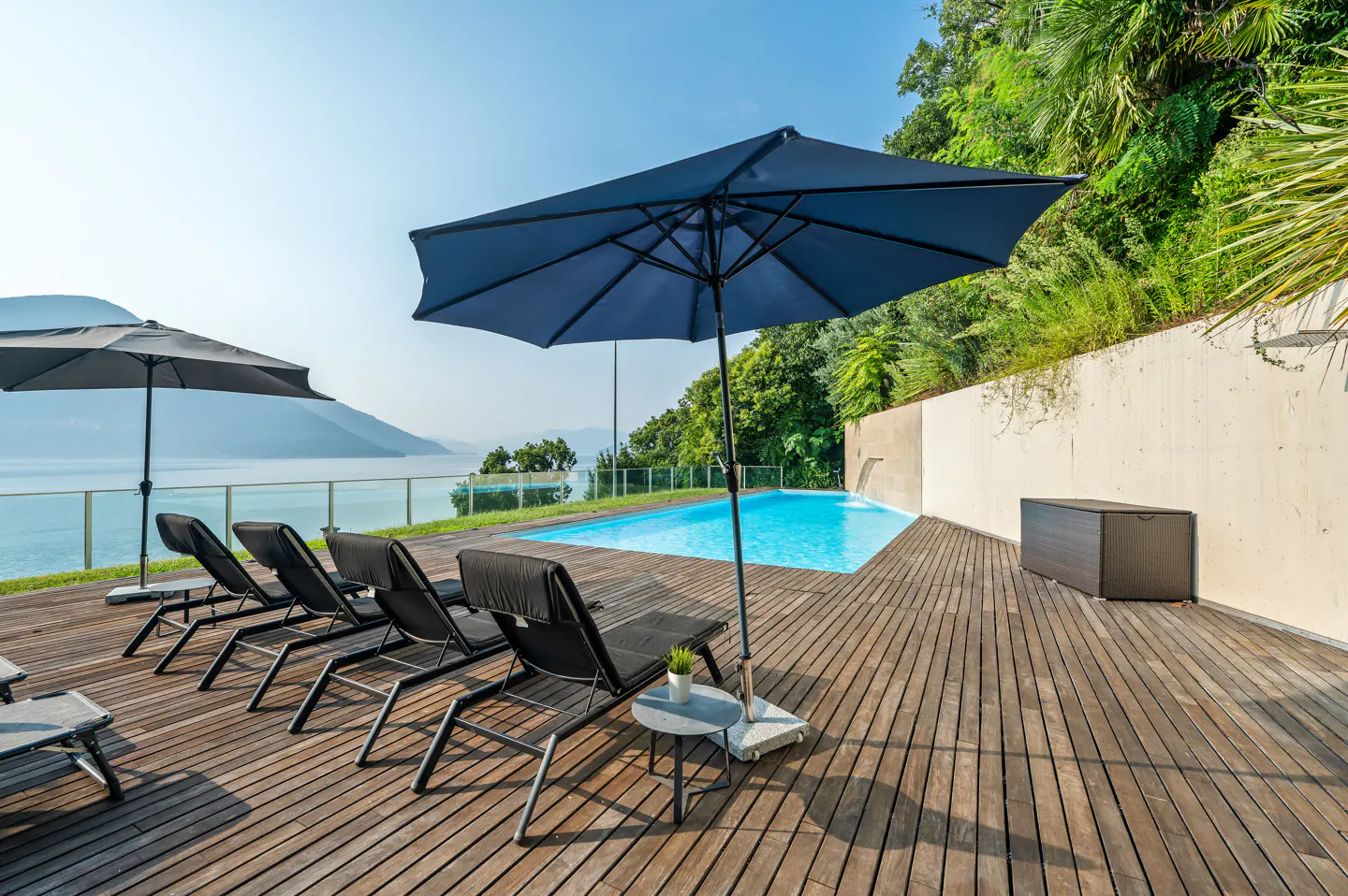 Outdoor pool area with lounge chairs, umbrellas, and a wooden deck overlooking a lake and mountains.