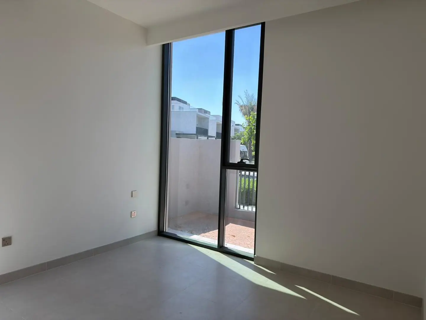 Empty room with white walls, light tile floors, and a tall window looking out to a sunny patio and neighboring houses.