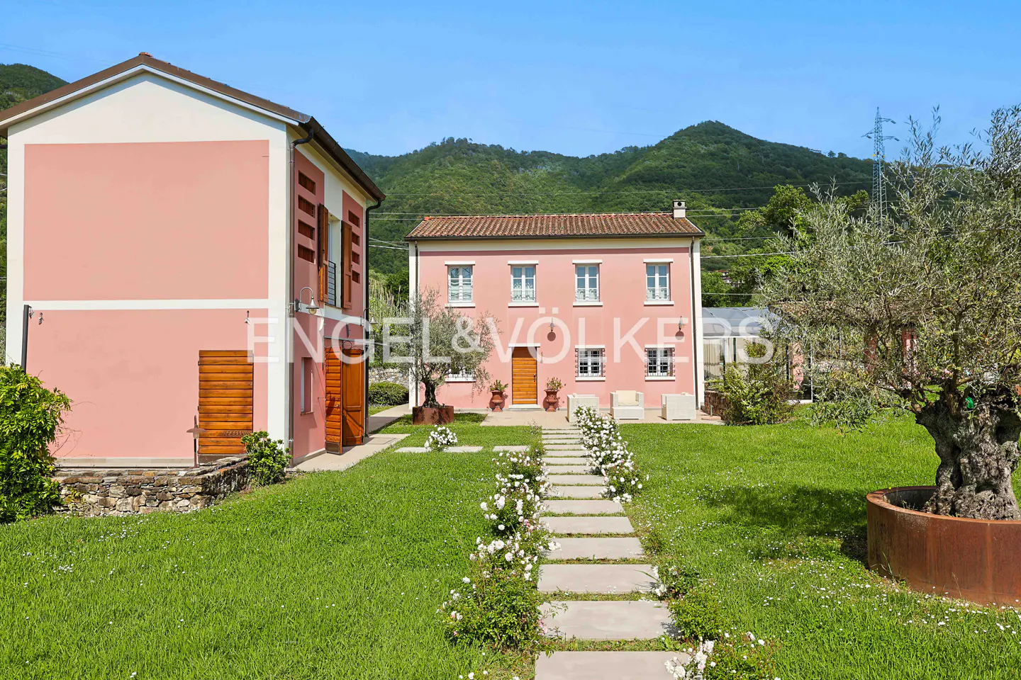 Exterior of a pink two-story house with a stone walkway and green lawn. Mountains are visible in the background.