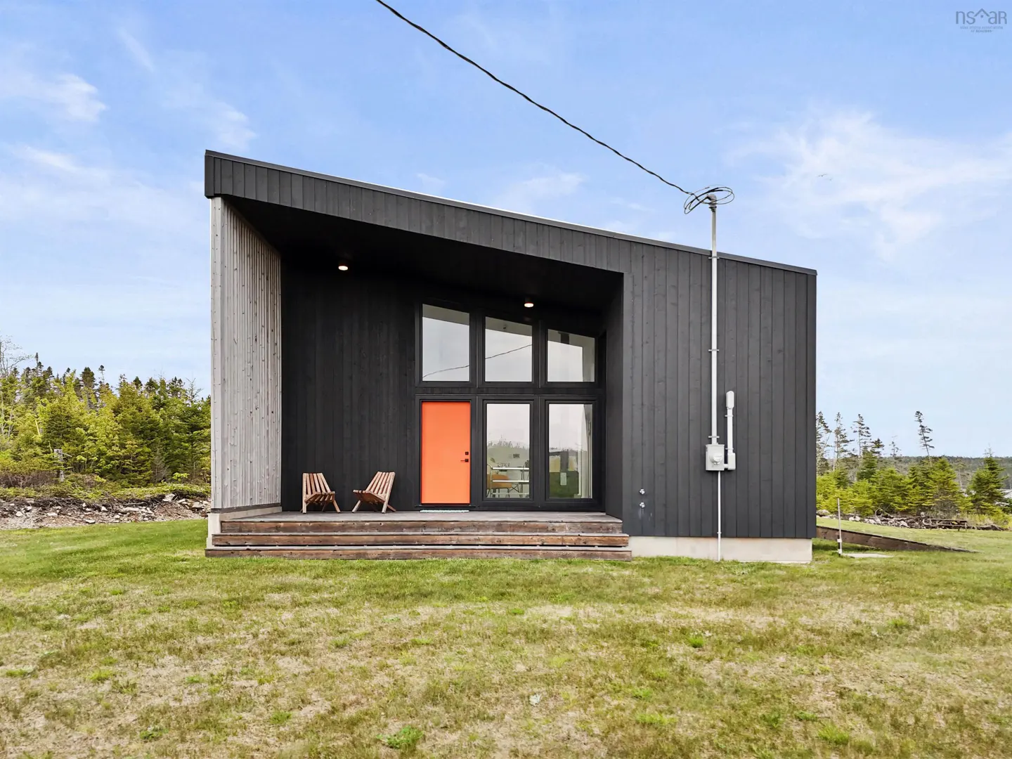 Modern black home with a slanted roof, bright orange front door, and two wooden chairs on a deck in a grassy yard.
