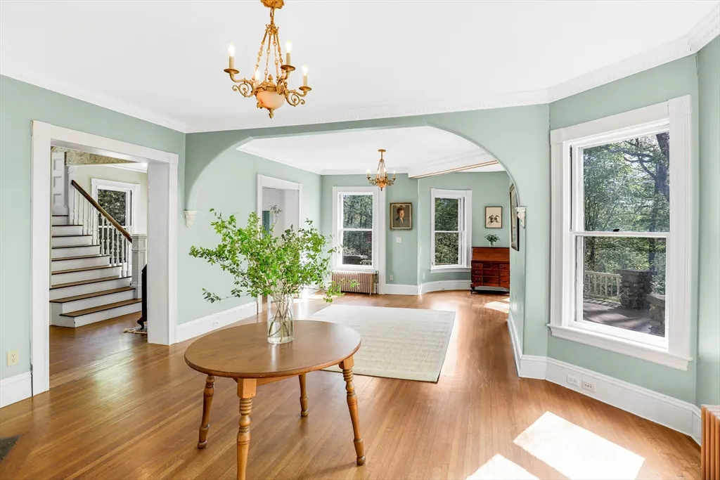 A bright, airy living room with light green walls, hardwood floors, and a staircase in the background. A round wooden table with a vase of greenery sits in the foreground.