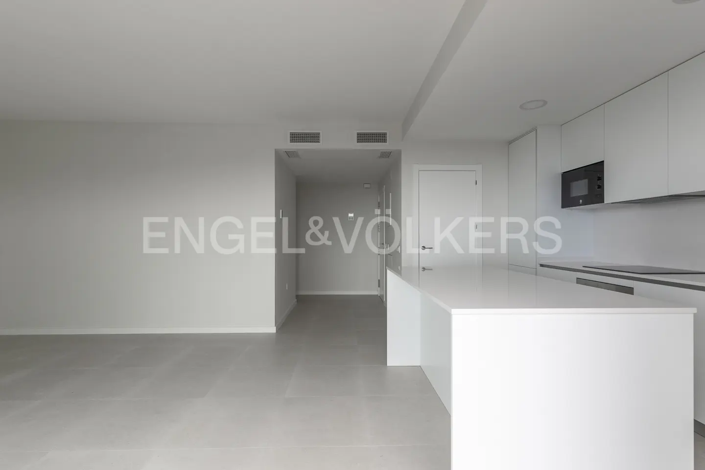 A bright, modern apartment with gray floors, white walls, and a white kitchen island. The Engel & Völkers logo is superimposed on the image.