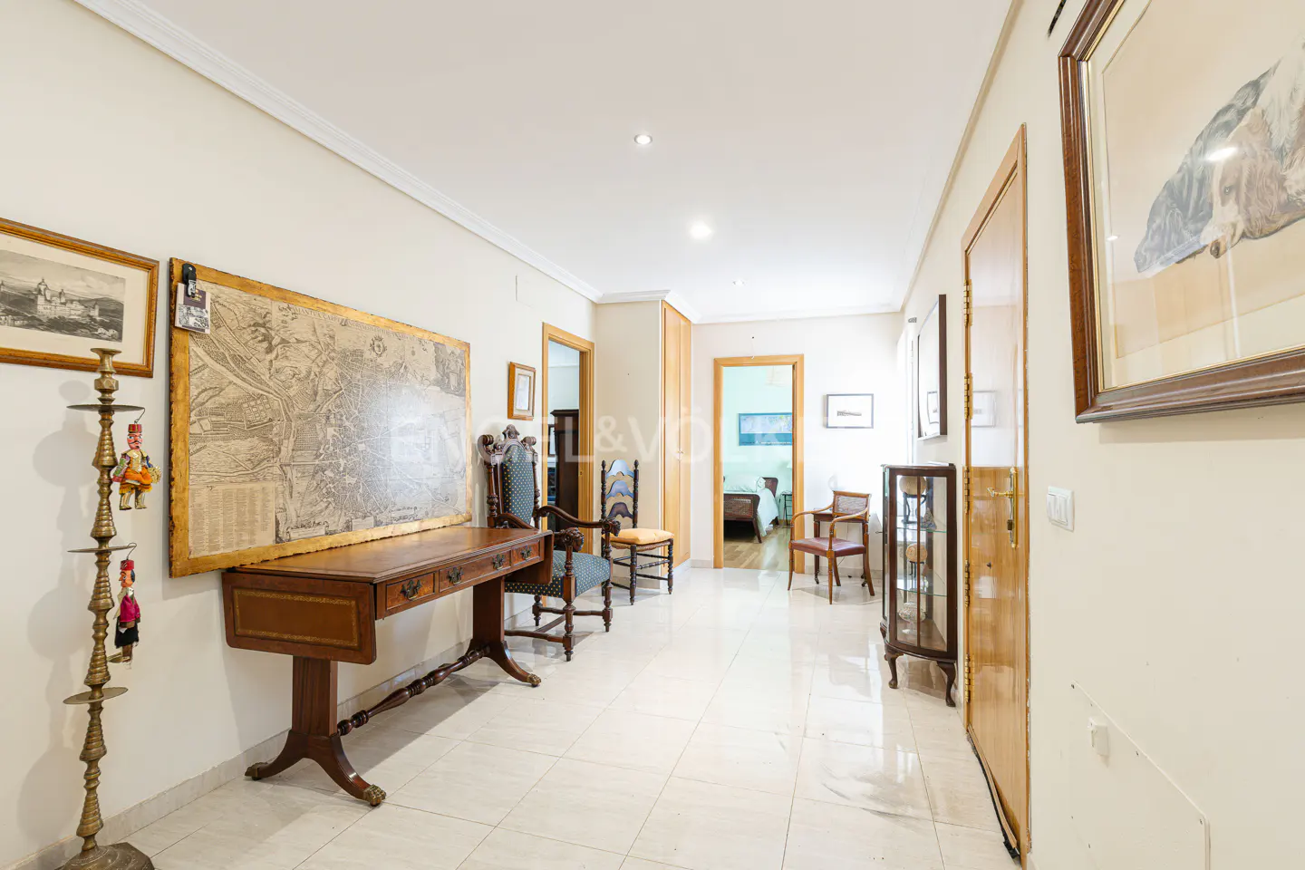 Hallway with cream walls, tiled floor, and antique furniture. A large map hangs above a wooden desk with chairs. Artwork adorns the walls.