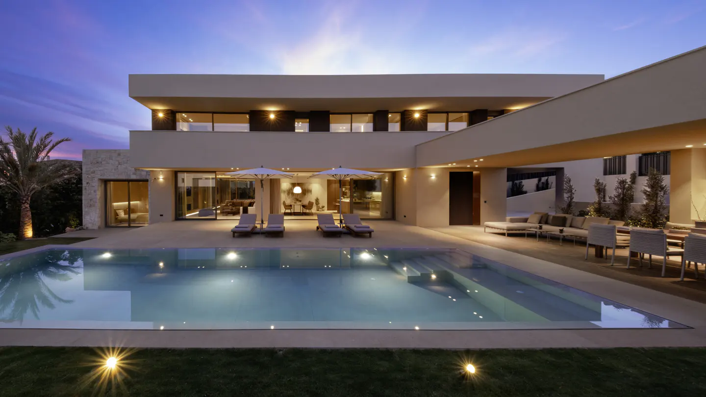 Modern beige house with a pool at dusk. Lounge chairs and umbrellas sit on the patio.
