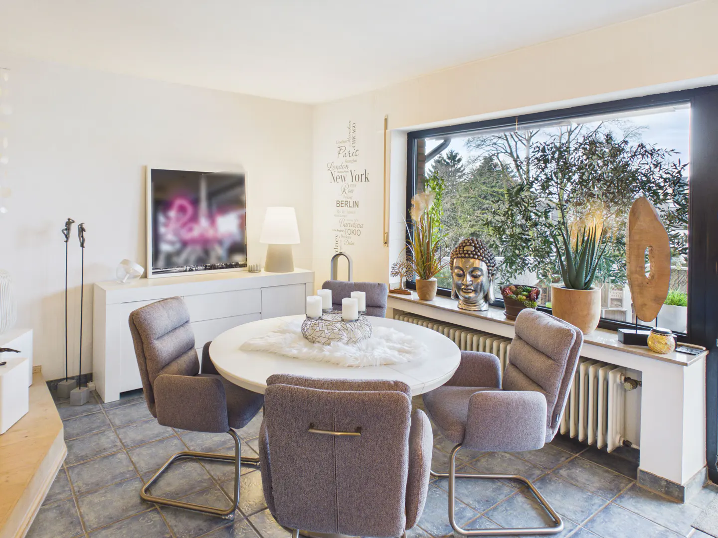 Bright dining room with a round table, gray chairs, and a large window with plants and a Buddha statue.