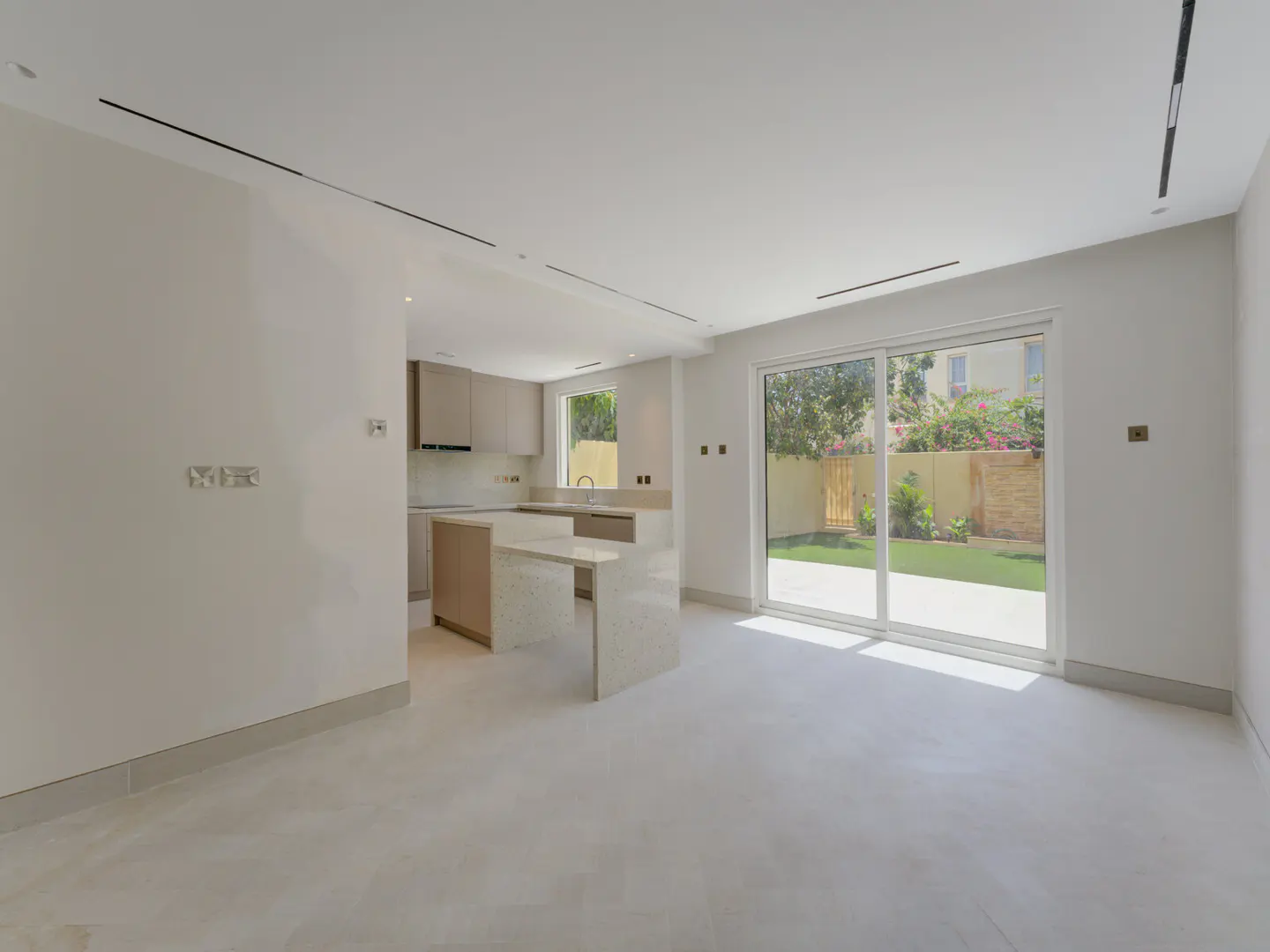 A bright, modern kitchen and living area with a large sliding glass door to a green backyard. Beige cabinets and a white island are visible.