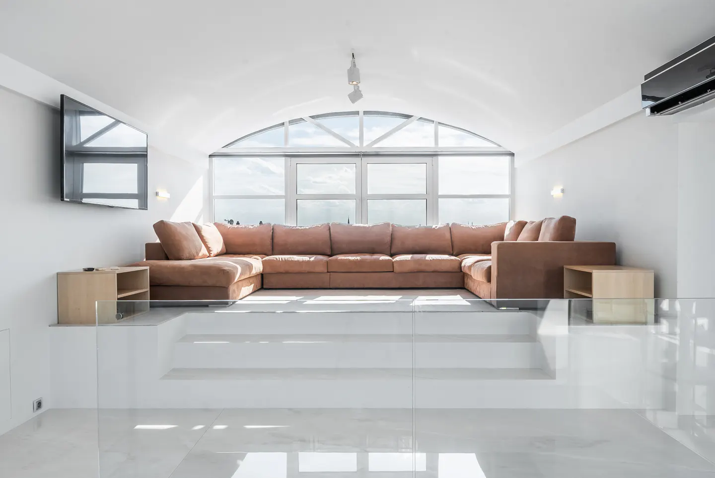 Bright, modern living room with a brown sectional sofa, white walls, and a large arched window. A TV is mounted on the wall.
