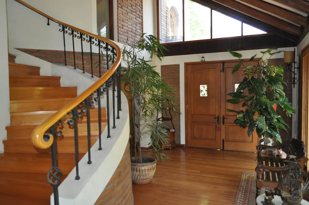 Interior view of a home's foyer with wood floors, a curved staircase with black iron railing, and double wood doors. Potted plants add greenery.