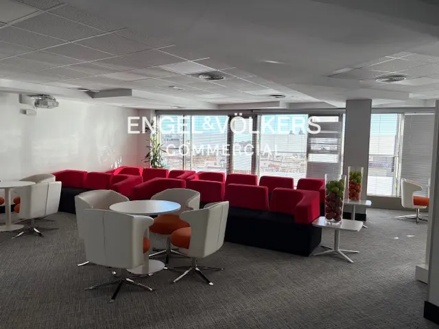 Office lounge area with red modular seating, white chairs, round tables, and large windows.