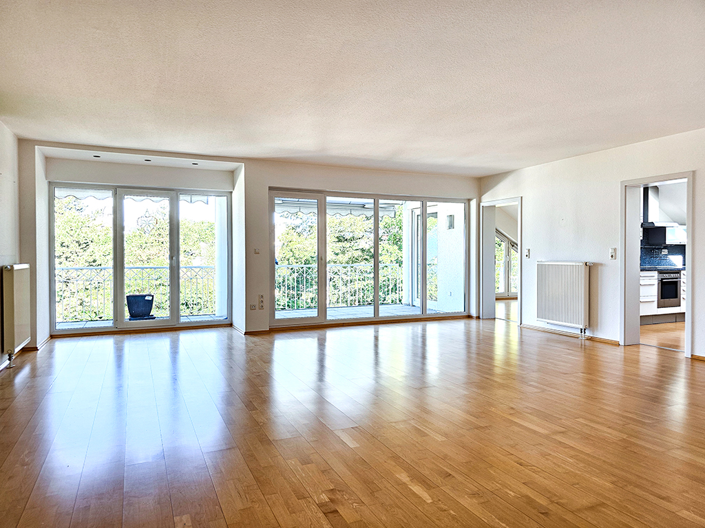 Spacious, empty living room with light wood floors, white walls, large windows, and a view of a balcony with greenery.