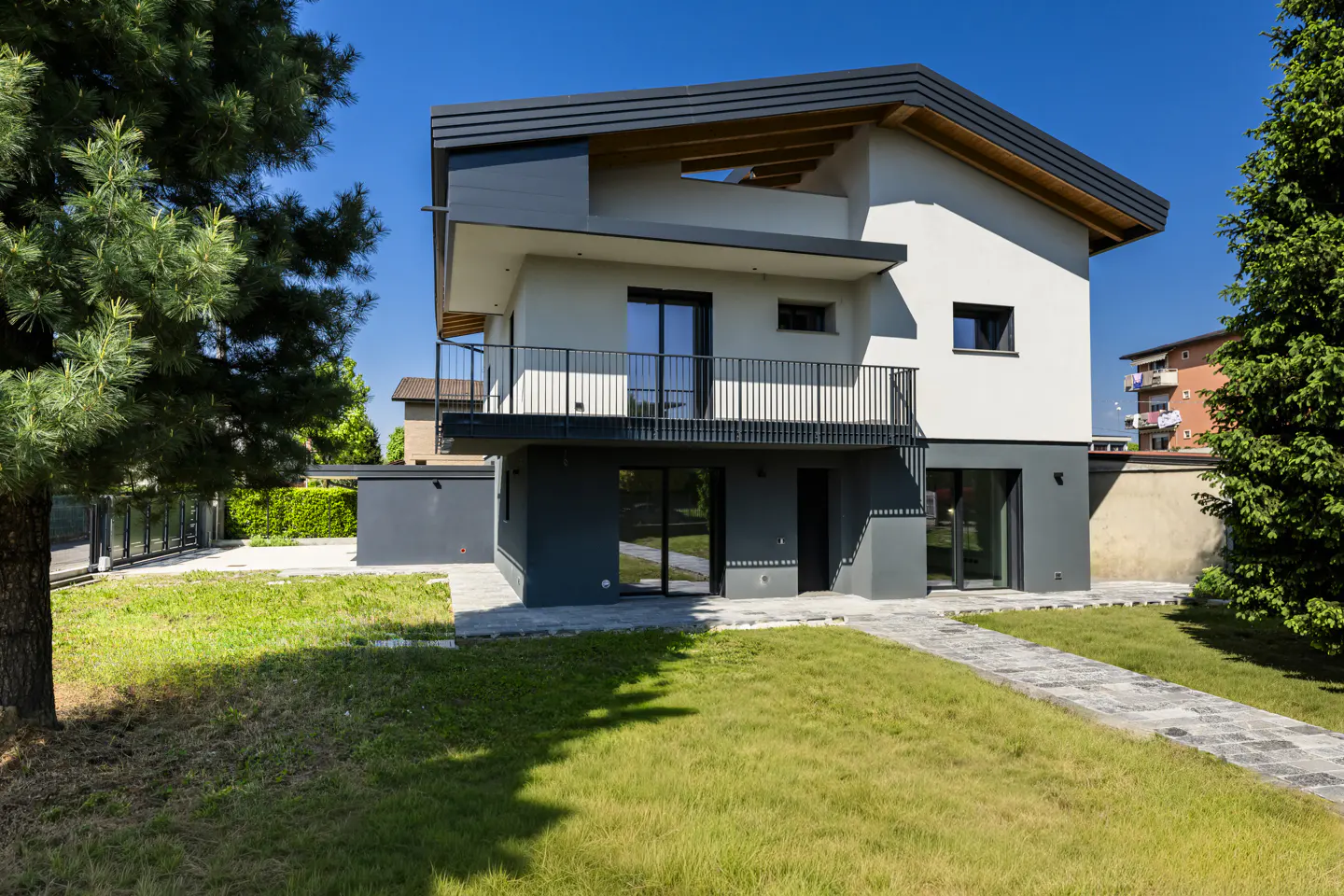 Two-story modern house with a gray and white exterior, black trim, and a small balcony, surrounded by a green lawn and trees.