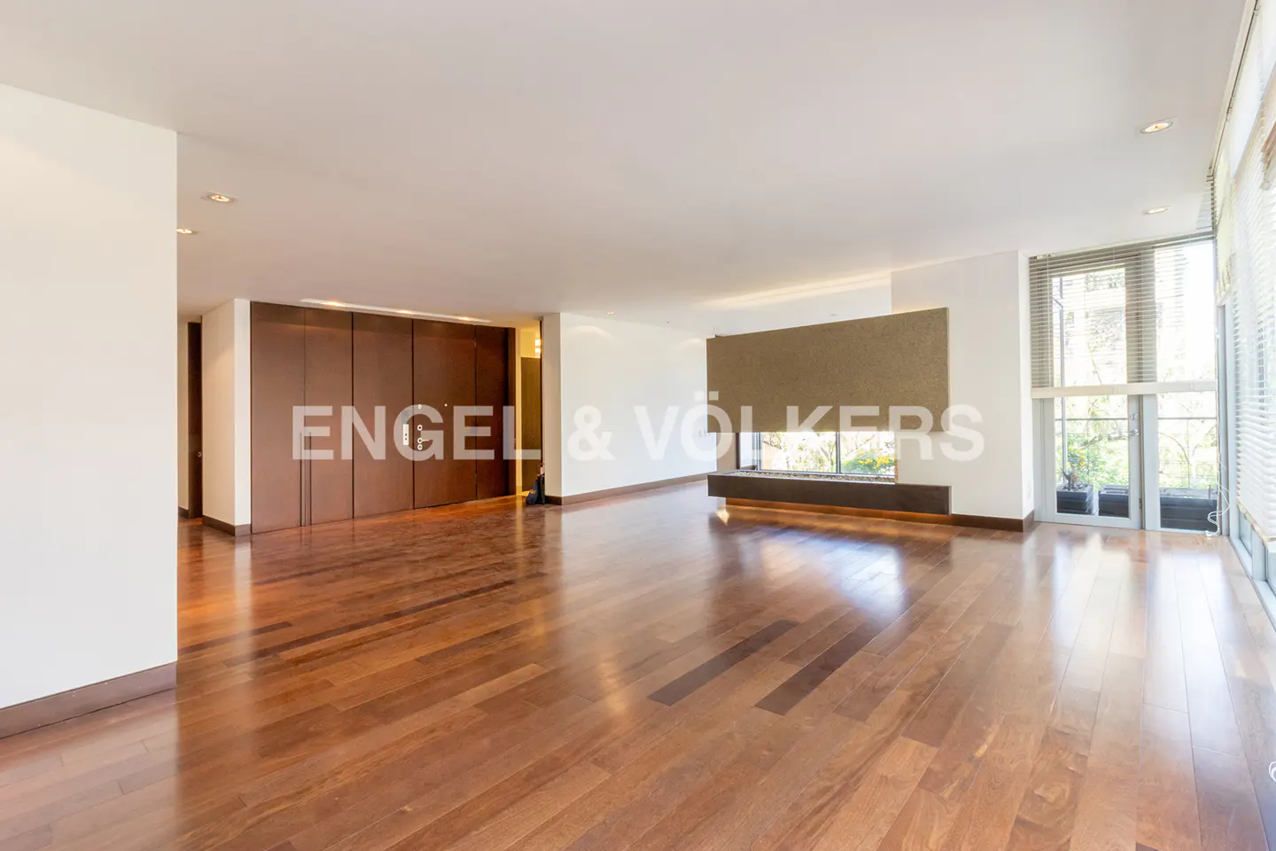 Spacious living room with hardwood floors, a fireplace, and large windows with blinds. Brown closet doors are on the left.