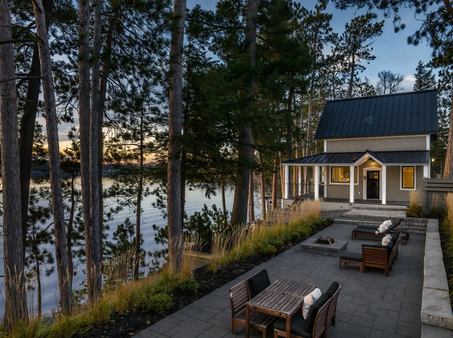 A tan house with a black roof sits near a lake, framed by tall trees. A stone patio features outdoor seating and a fire pit.