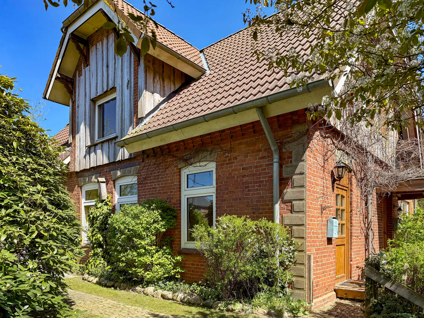 Exterior view of a two-story brick house with a brown tiled roof and white-framed windows, surrounded by green bushes.