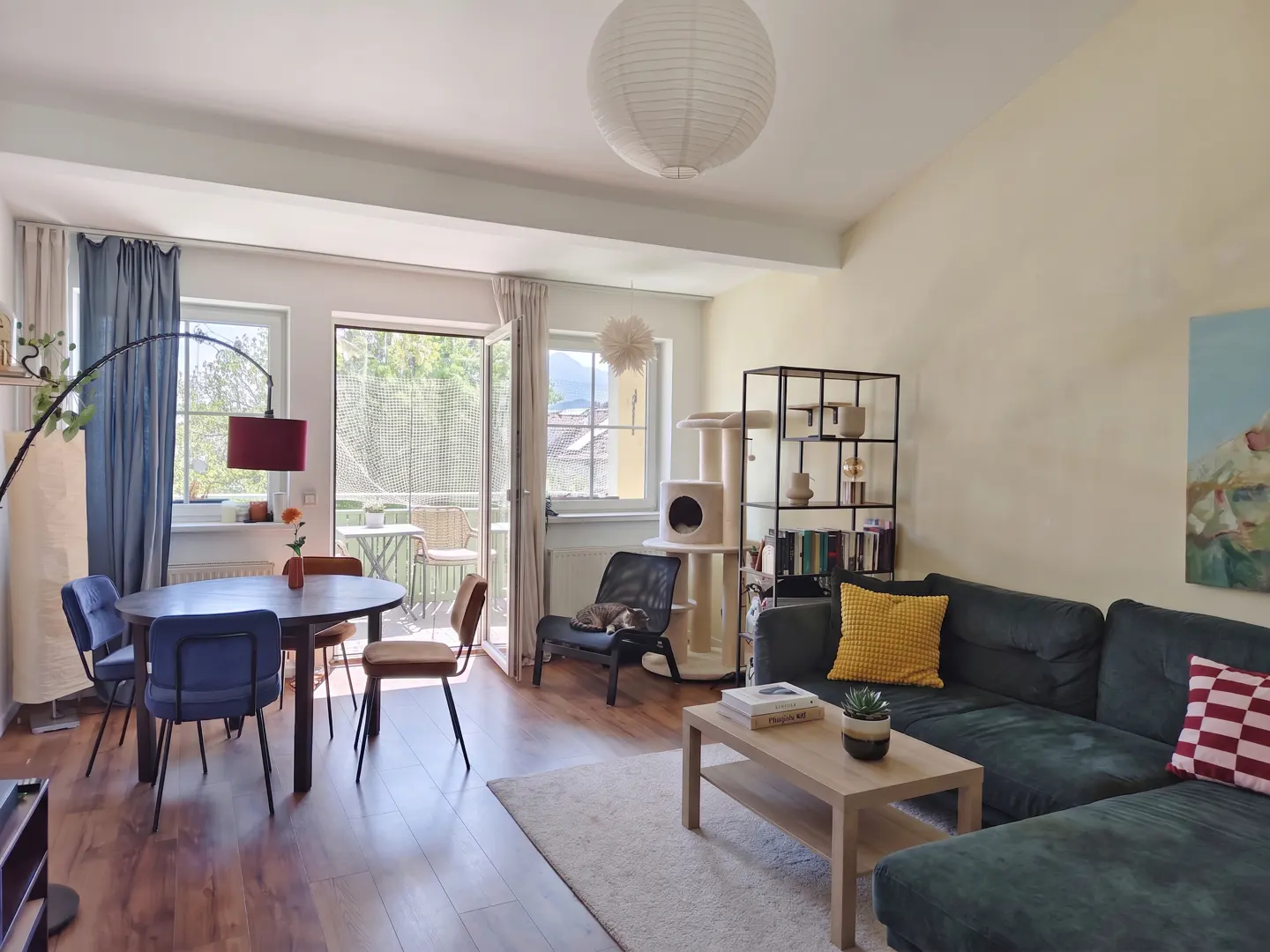 Bright living room with wood floors, a dark green sectional sofa, a dining table with blue chairs, and a cat sleeping in a black chair. Balcony in background.