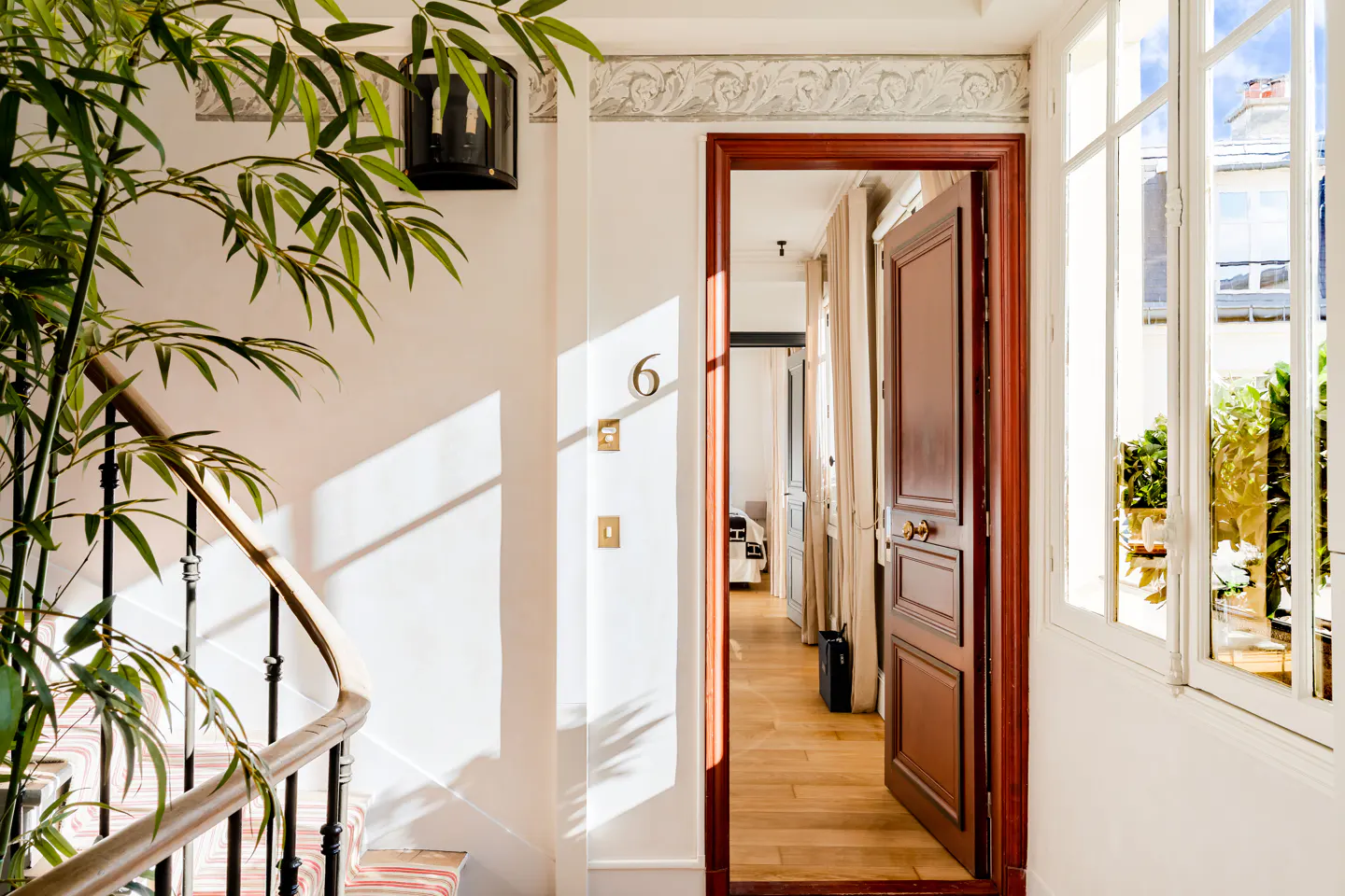 Bright hallway with a potted plant, a staircase with a wooden railing, and an open door leading to a sunlit room.