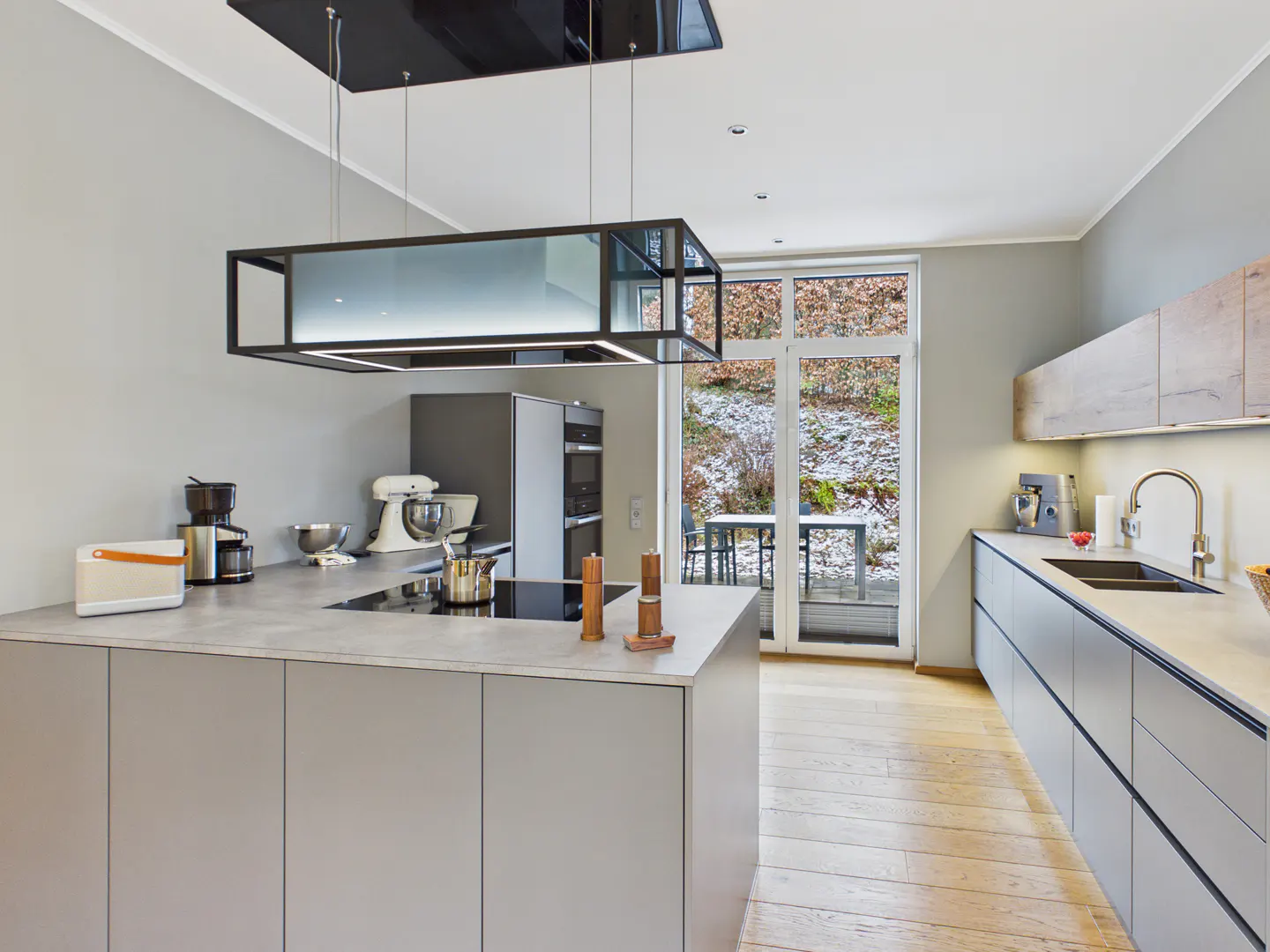 Modern kitchen with gray cabinets, light wood floors, and a black rectangular light fixture. A glass door leads to a snowy outdoor patio.