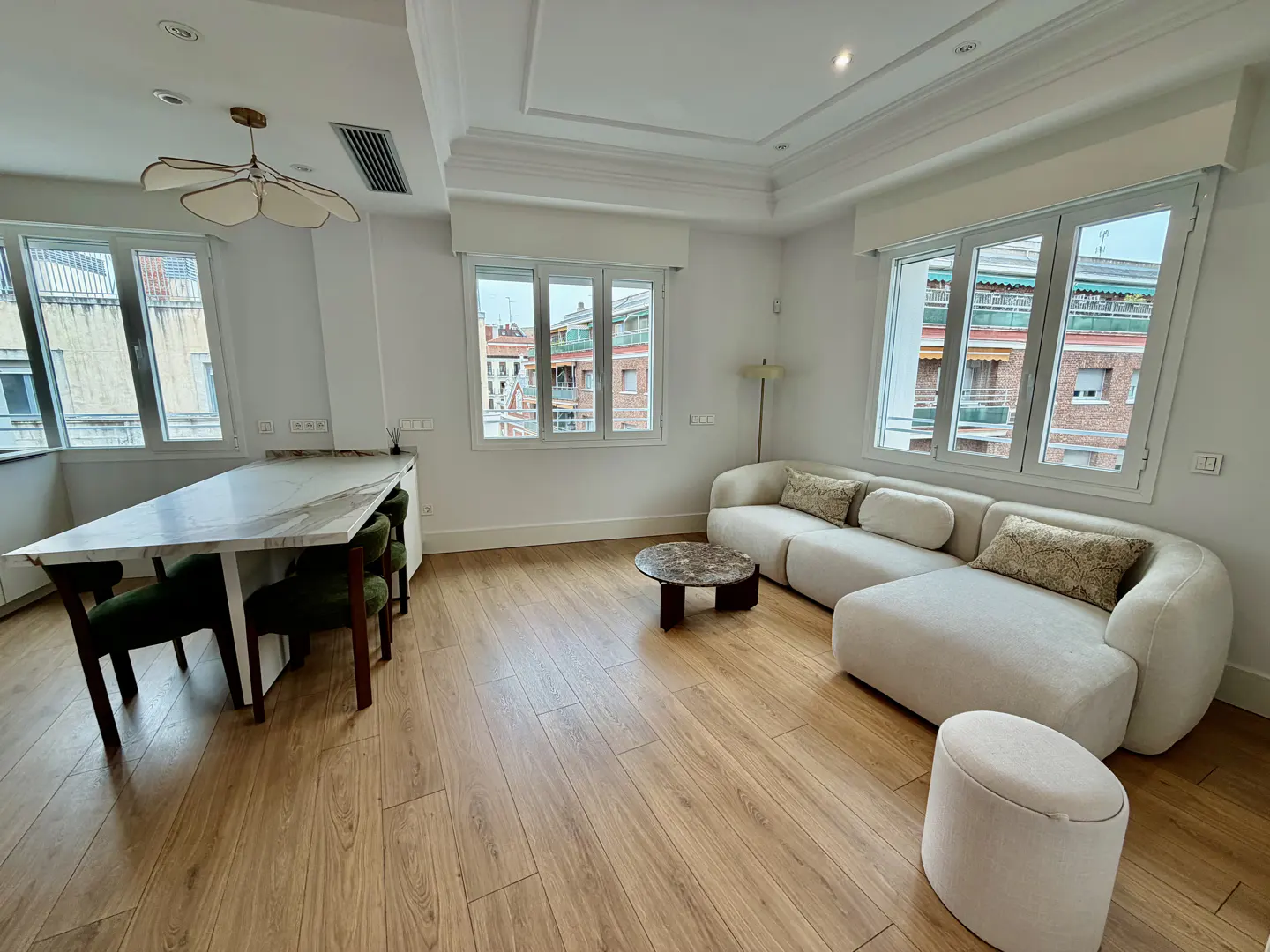 Bright living room with wood floors, white walls, and large windows. A white sofa, marble table, and green chairs furnish the space.