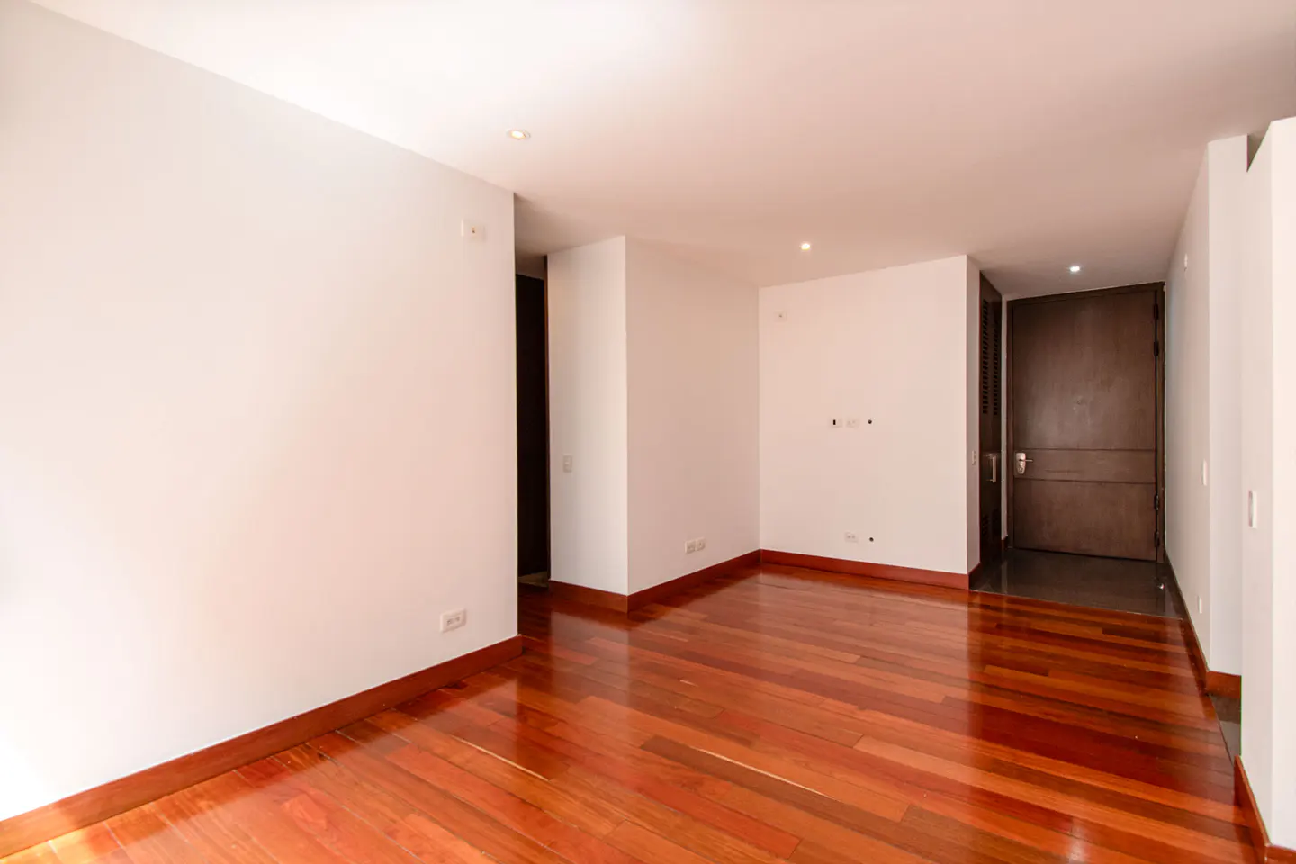 Bright, empty room with white walls and a glossy, reddish-brown hardwood floor. A dark wood door is visible in the background.
