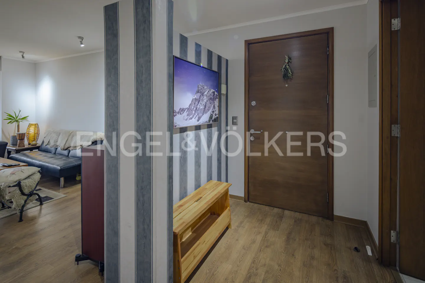 Interior view of a home with a wooden door, striped wallpaper, and a TV displaying a mountain scene. A wooden bench sits below the TV.