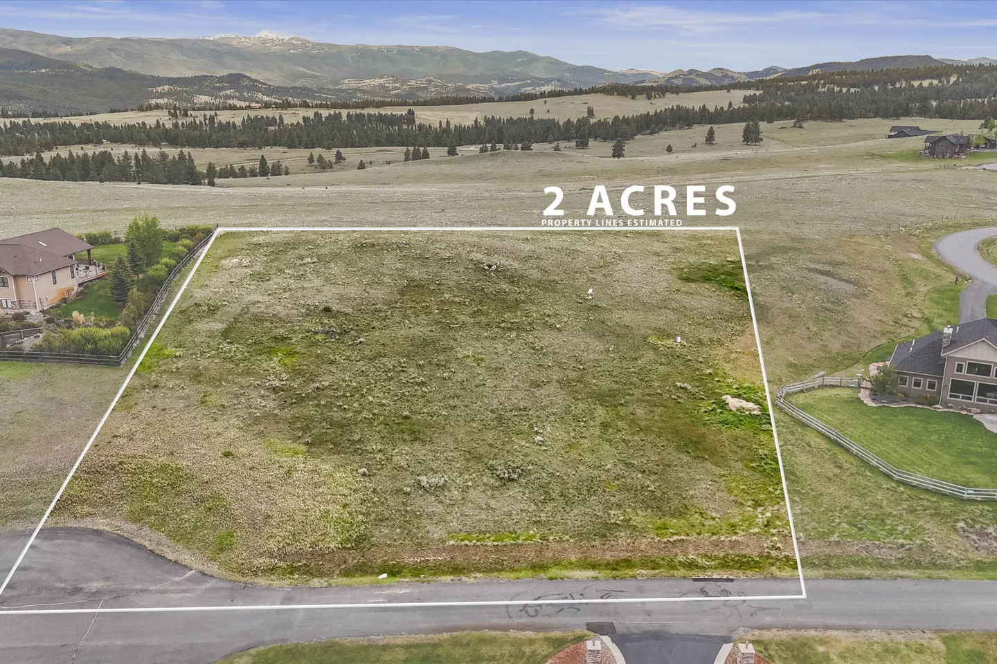 Aerial view of a 2-acre grassy lot outlined in white, with mountains and trees in the background. Houses are visible on either side of the lot.