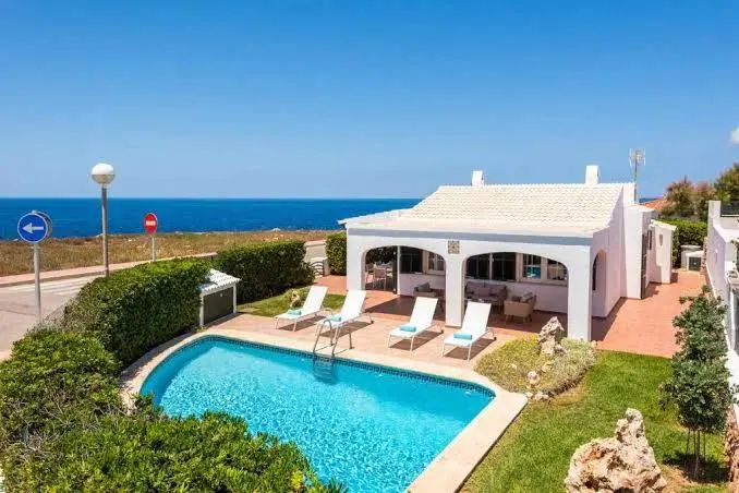 Aerial view of a white house with a pool, lounge chairs, and ocean view under a clear blue sky.