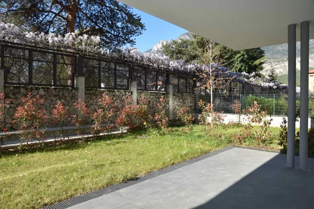 A patio with gray tiles overlooks a green lawn, a stone wall with a black metal fence, and mountains in the background.
