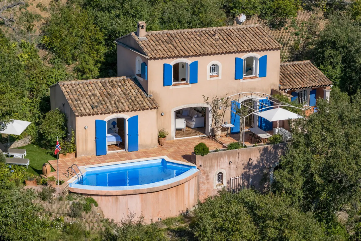 Aerial view of a peach-colored villa with blue shutters, a terracotta roof, and a blue swimming pool surrounded by lush green trees.
