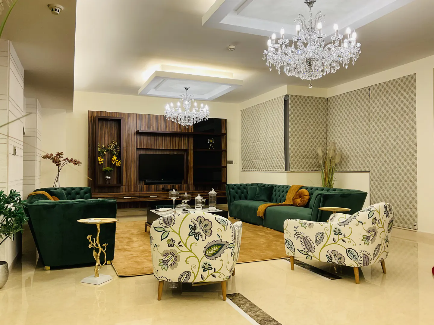 A living room with green sofas, floral chairs, a TV, and crystal chandeliers.