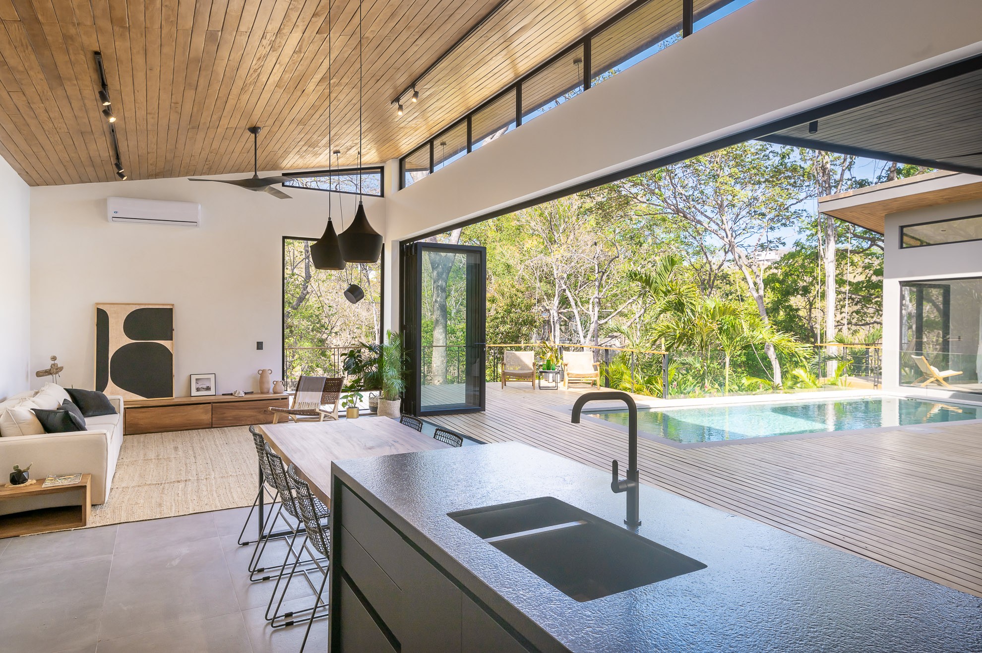 Modern open-plan kitchen and living area with a wooden ceiling, folding glass doors, and a deck with a swimming pool.