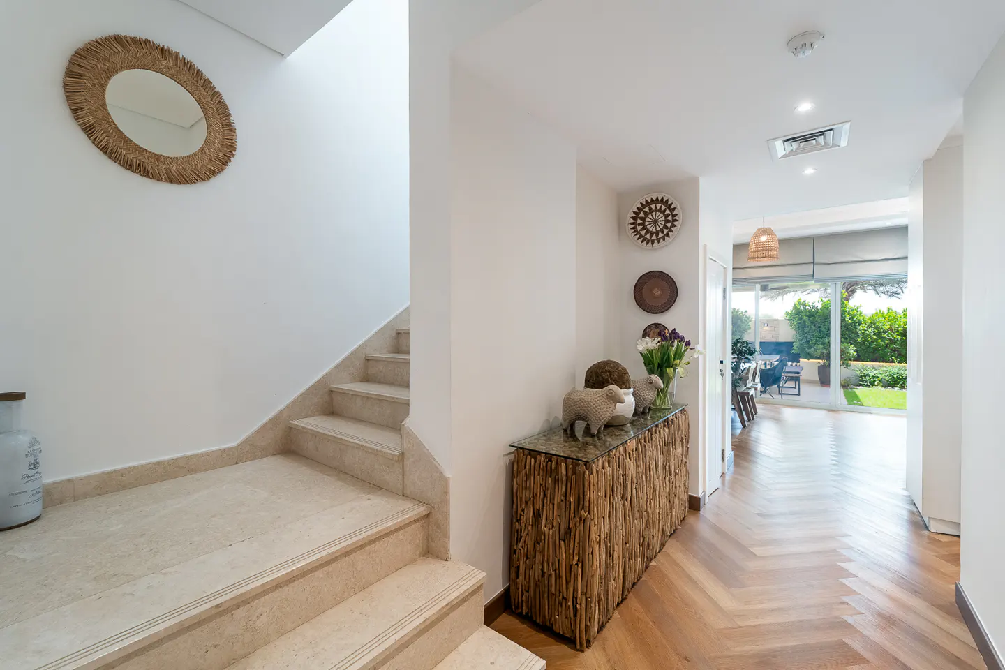 Hallway with beige stairs, a round mirror, and a wooden console table with decor. A view to a garden is visible through glass doors.