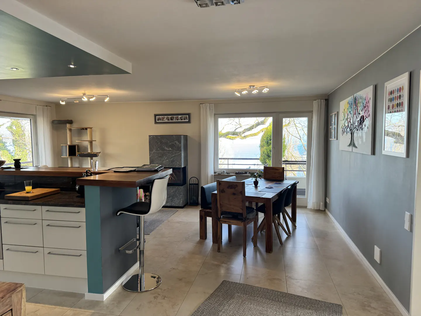 Open-concept living space with kitchen island, dining table, and gray accent wall with artwork. Large windows offer a view.