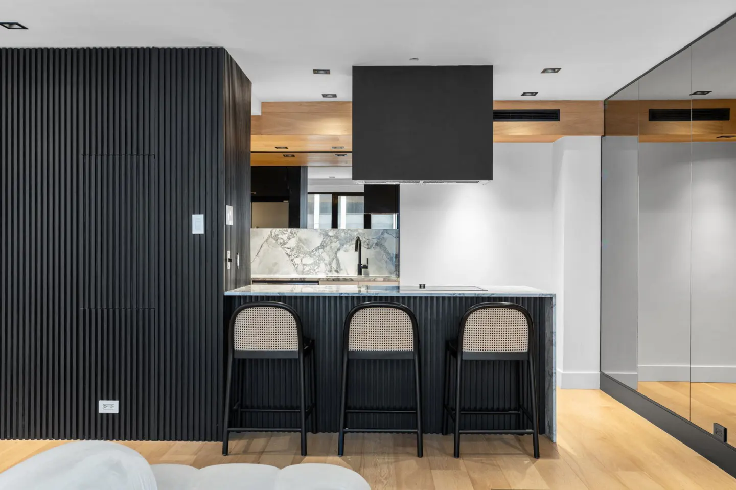 Modern kitchen with black cabinetry, marble countertops, and three black bar stools with woven backs. A black range hood hangs above the island.